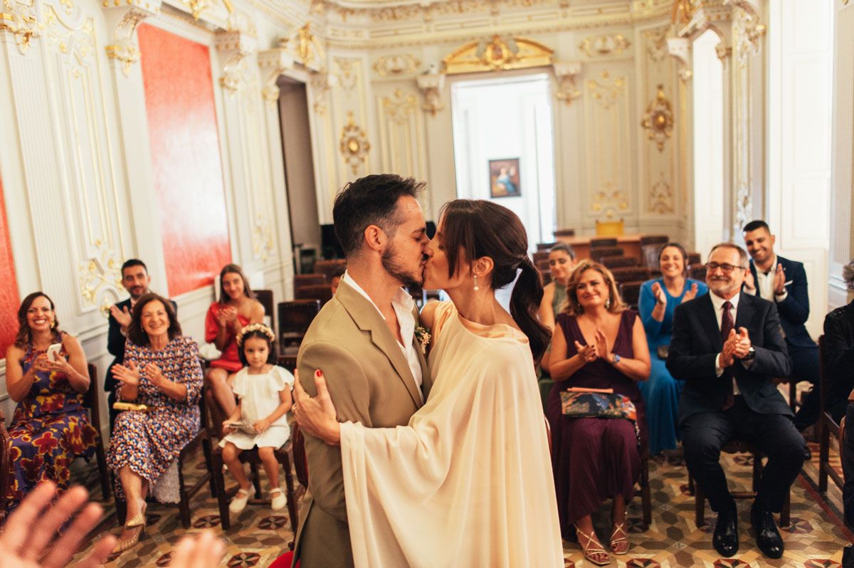 Novios besándose justo después de la ceremonia civil, rodeados de sus invitados en el salón del ayuntamiento