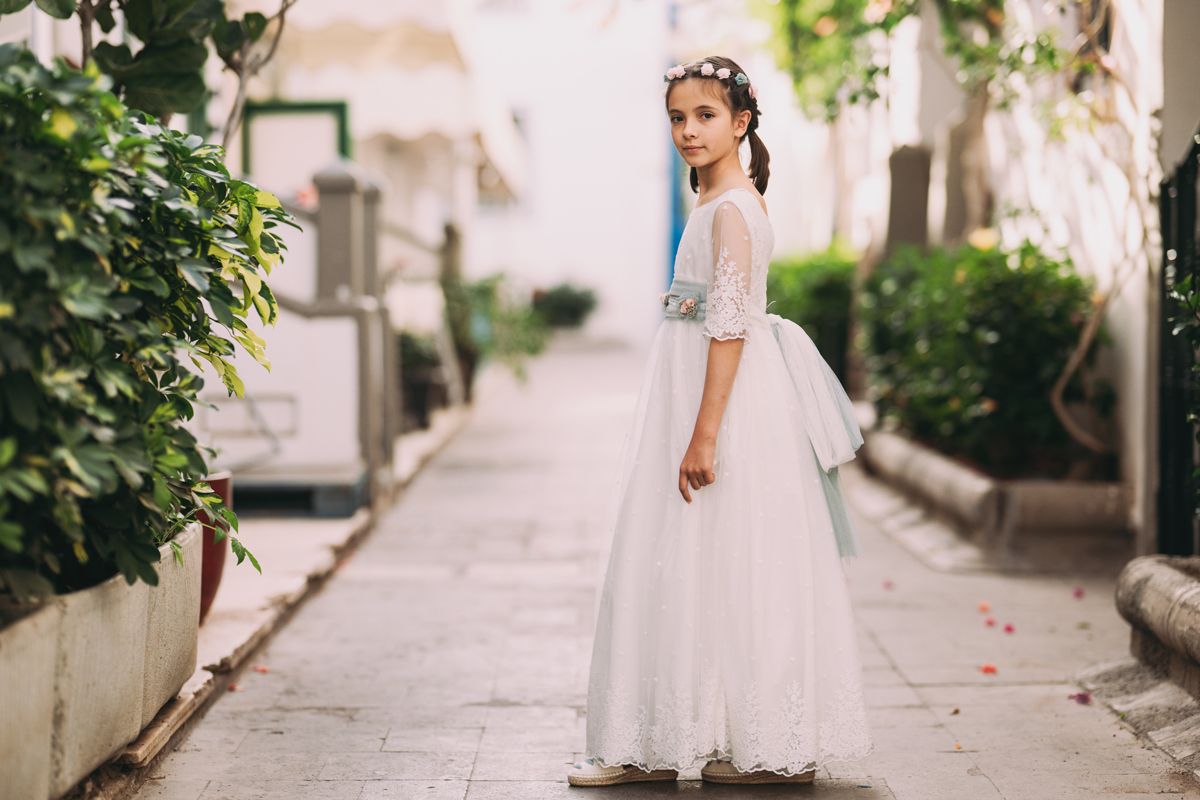 Niña con vestido de comunión de encaje y corona de flores paseando por una calle empedrada de Madrid