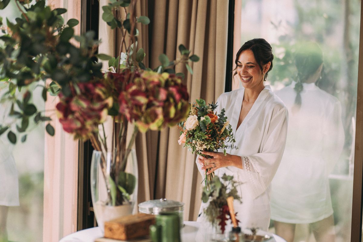 Novia preparandose antes de la boda, sonriendo con el ramo natural en la mano